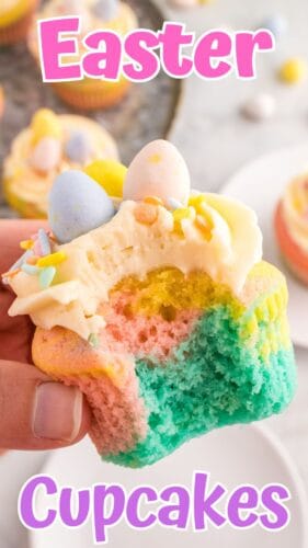 A pastel cupcake is held in a hand, revealing colorful layers. It has buttercream frosting, sprinkles, and mini eggs. The background shows more cupcakes. A pink and purple text overlay reads "Easter Cupcakes."