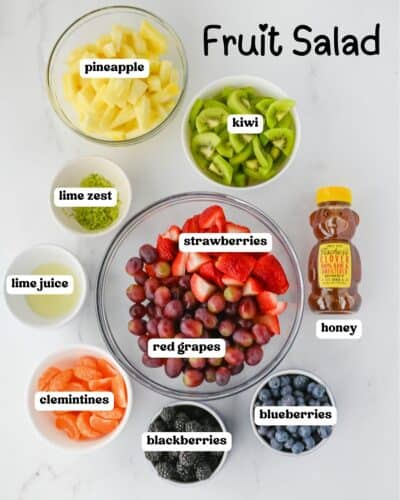Flat lay photo showing prepped ingredients for fruit salad in small bowls. Includes labeled items: pineapple, kiwi, strawberries, red grapes, clementines, blackberries, blueberries, lime zest, lime juice, and a honey bottle.